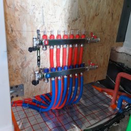 HYDRO-MANN - Rozdzielacz ogrzewania podłogowego z czerwonymi i niebieskimi rurami na folii izolacyjnej, zamontowany na ścianie z płyty OSB. Widok na instalację w trakcie montażu.