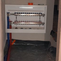 HYDRO-MANN - Rozdzielacz ogrzewania podłogowego w białej szafce, z niebieskimi i czerwonymi rurami, w surowym wnętrzu. Na szafce pomarańczowa taśma.