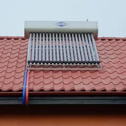 Kolektor słoneczny Sanko z rurami na czerwonym dachu. Widoczne mocowania i instalacja hydrauliczna. Szare niebo w tle.