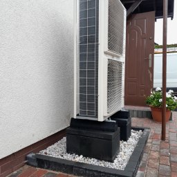 Biała pompa ciepła typu split, ustawiona na czarnych betonowych blokach, otoczona białym żwirem i kostką brukową, na tle białej ściany budynku i brązowych drzwi.