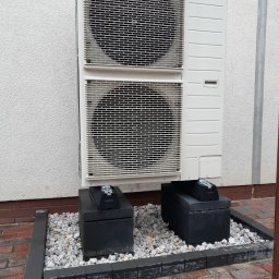 HYDRO-MANN - Biała, dwuczęściowa pompa ciepła Panasonic zamontowana na betonowych blokach, otoczona kamieniami i obrzeżem z kostki, przy ścianie budynku.