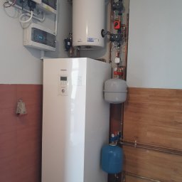 HYDRO-MANN - Biała pompa ciepła Panasonic Aquarea w kotłowni, z widocznymi elementami instalacji, zbiornikiem i orurowaniem. Na ścianie panele sterowania i zabezpieczeń.