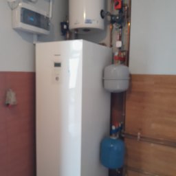 HYDRO-MANN - Biała pompa ciepła Panasonic z zbiornikiem, widoczne rury i zawory. Elementy instalacji grzewczej na tle ściany w dwóch kolorach. Ujęcie z bliska, lekko zamazane.