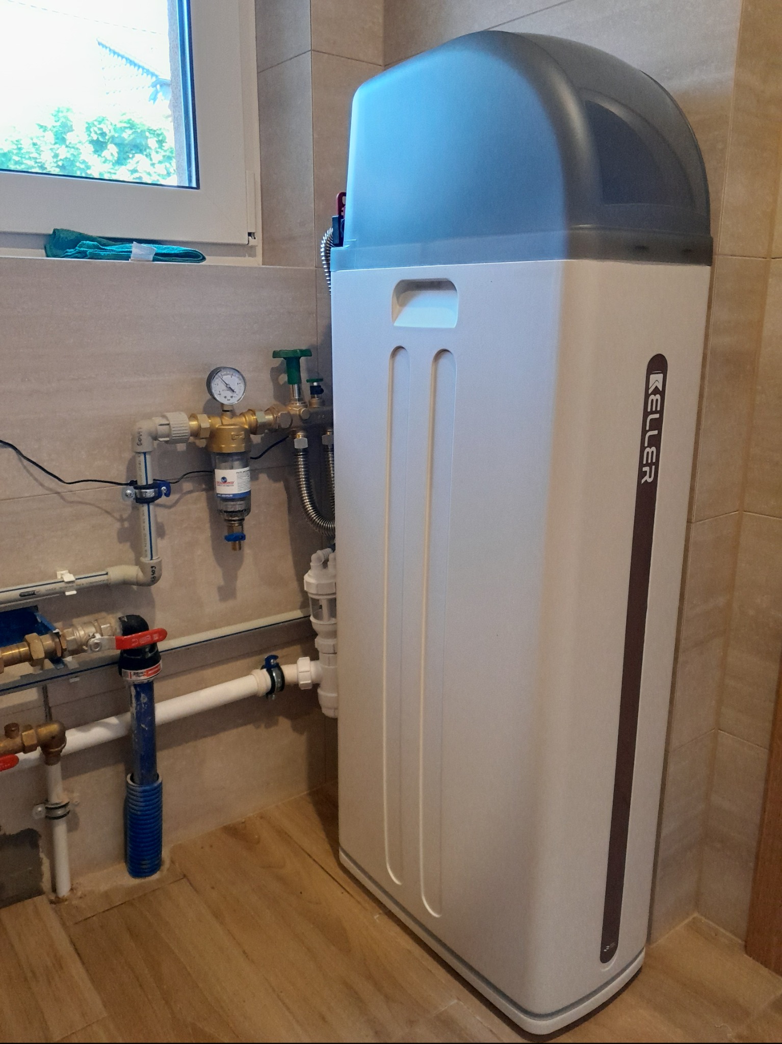 Filtr do wody marki Keller zainstalowany w pomieszczeniu z beżowymi płytkami i oknem. Widoczne rury i zawory. Filtr stoi na podłodze imitującej drewno.