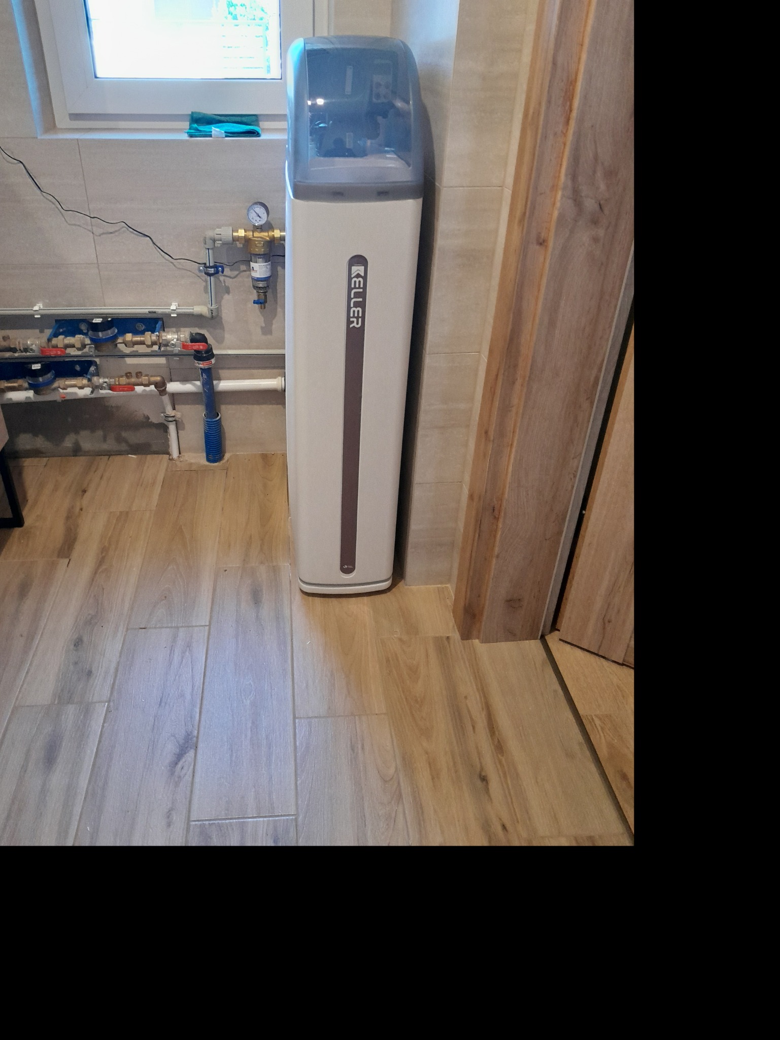 Biały zmiękczacz wody Keller w łazience z drewnopodobną podłogą i instalacją hydrauliczną w tle, widok z góry.
