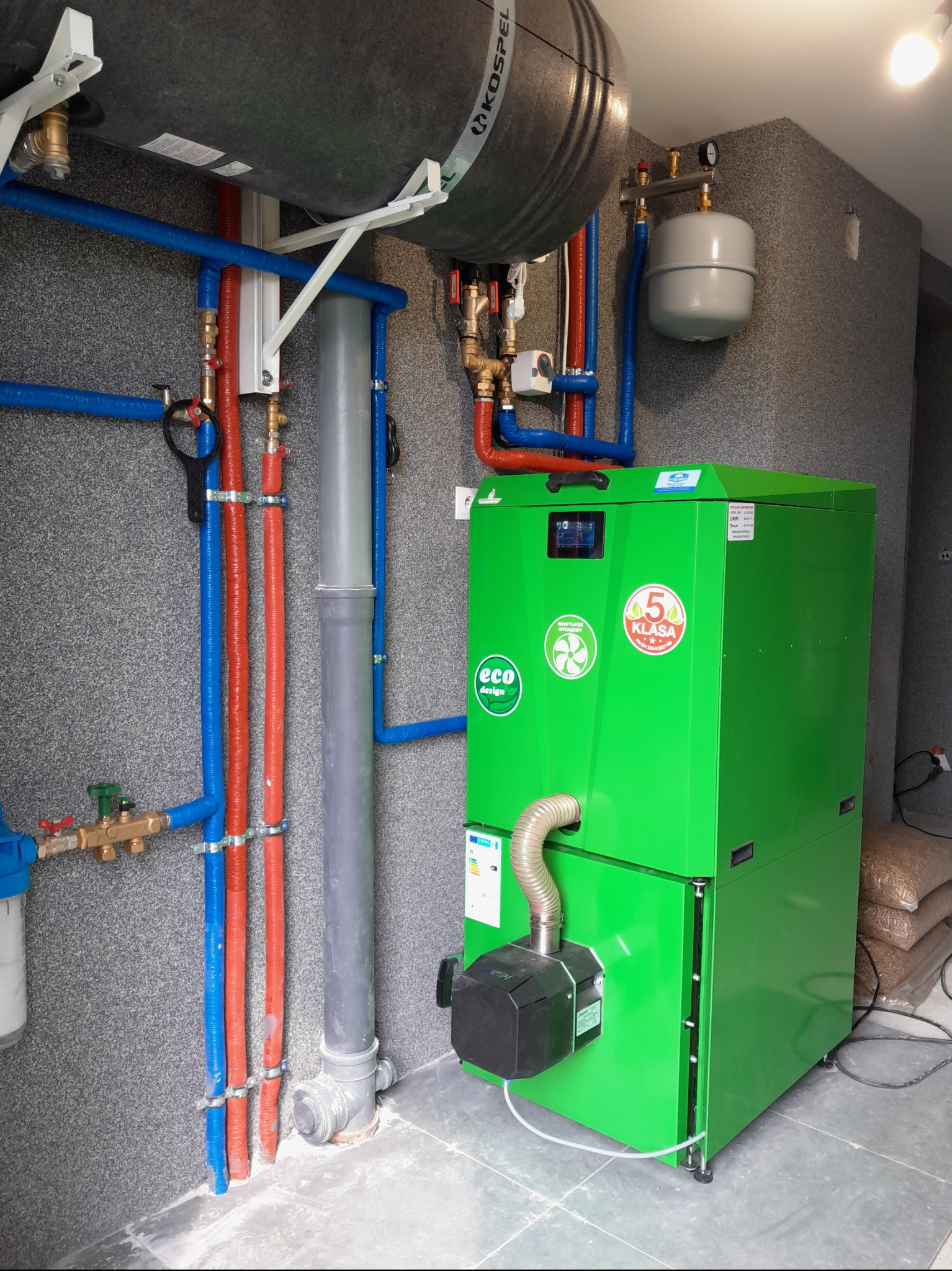 Zielony piec na pellet z instalacją CO, zbiornik Kospel i rurami w kolorze czerwonym i niebieskim na tle ściany z szarego kamienia.