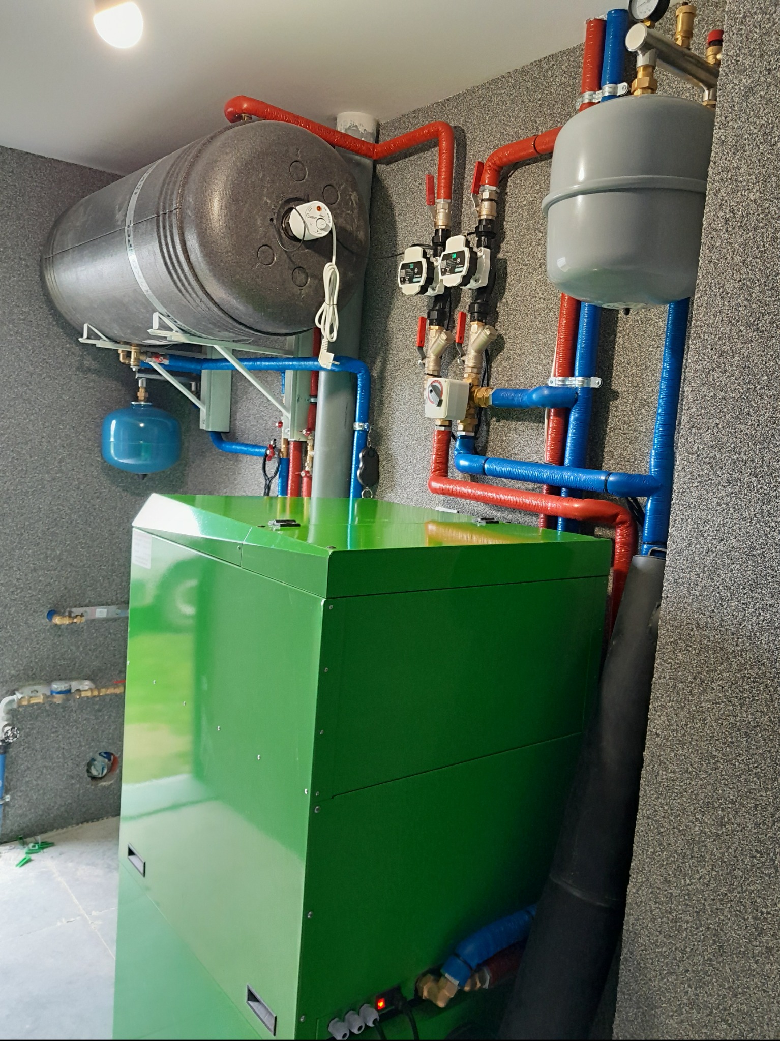 Zielony piec grzewczy z instalacją: zbiornik szary, rury czerwone i niebieskie. Kompaktowy system grzewczy w pomieszczeniu technicznym, widok z dołu.
