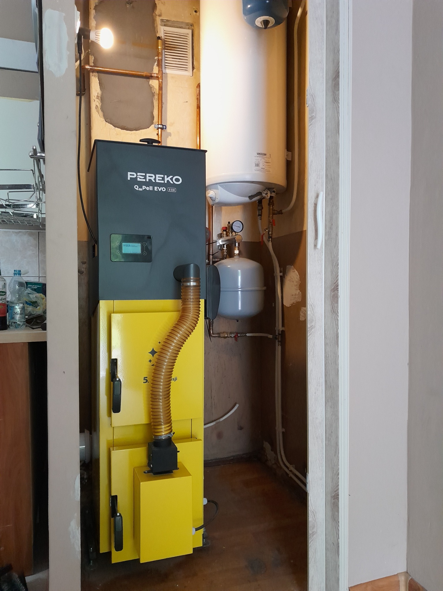 Zamontowany, nowoczesny piec Pereko QmPell EVO 8 kW z żółtym podajnikiem, obok biały zasobnik ciepłej wody i szary zbiornik wyrównawczy w kotłowni.
