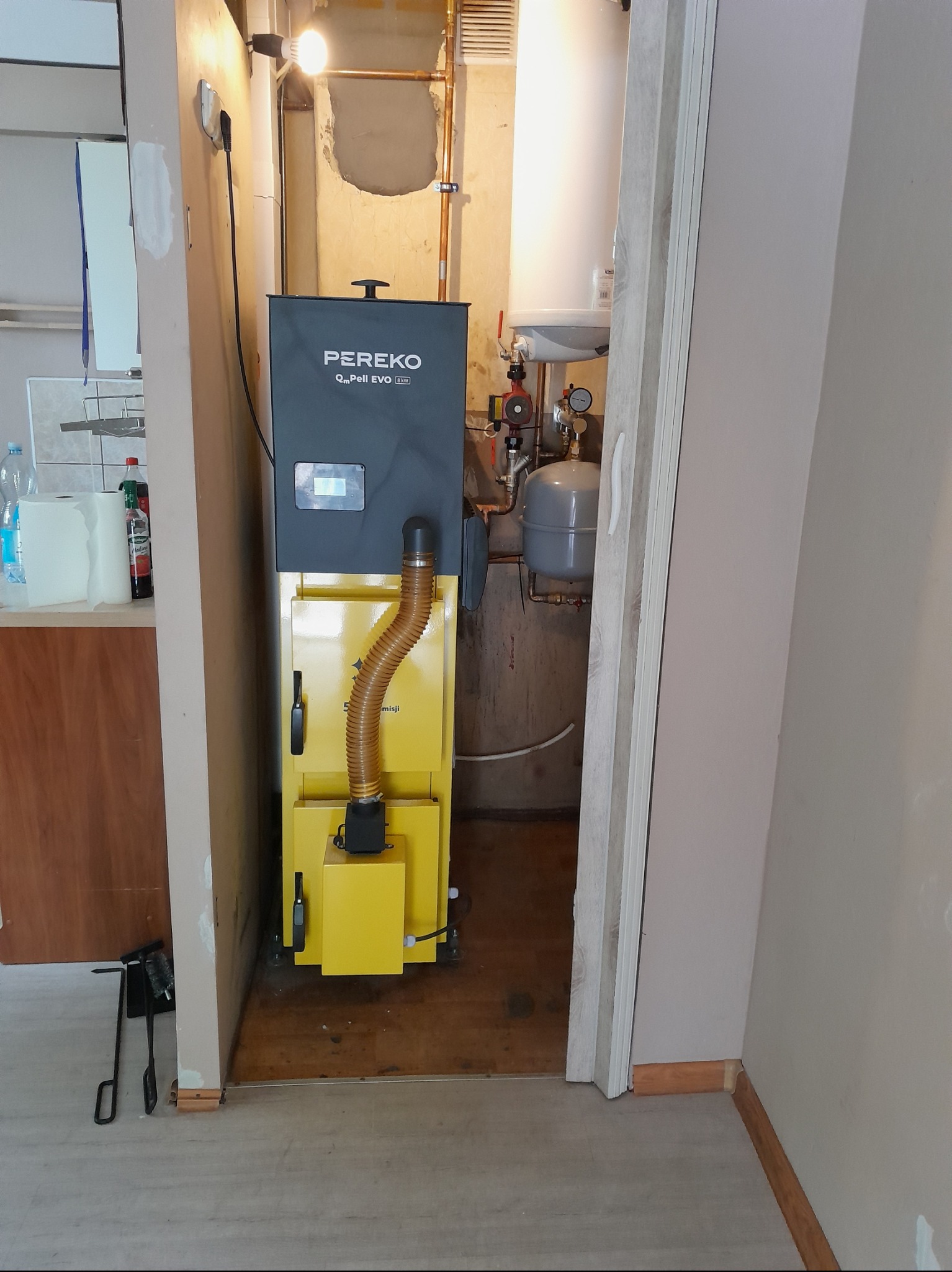 Żółto-szary piec na pellet Pereko Q-Pell EVO BW w kotłowni. Widoczne rury miedziane, zbiornik wyrównawczy i akcesoria do czyszczenia pieca obok drzwi.