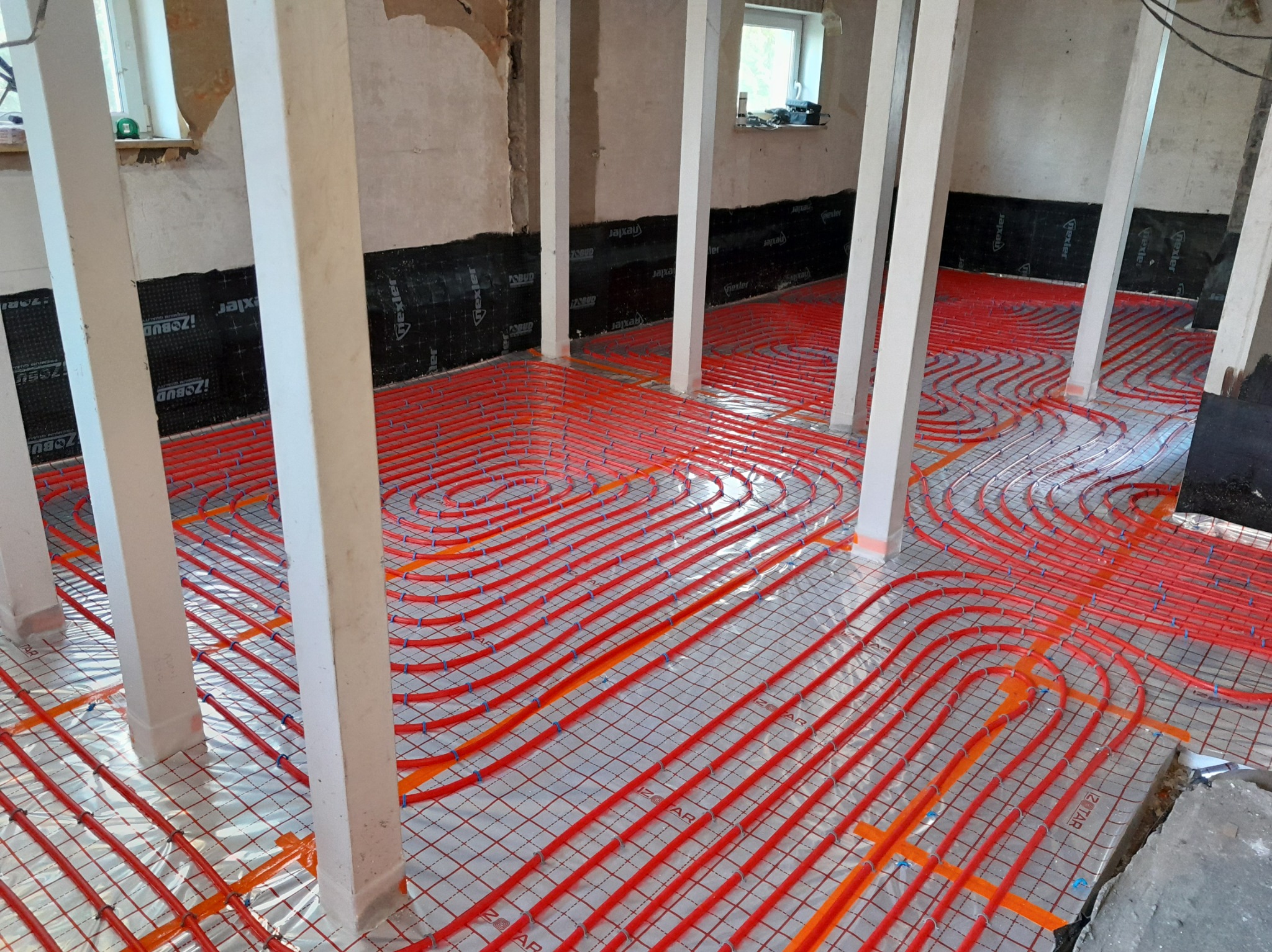 Czerwone rury ogrzewania podłogowego ułożone na folii izolacyjnej w pomieszczeniu z białymi filarami. Widoczne czarne maty izolacyjne na ścianach.