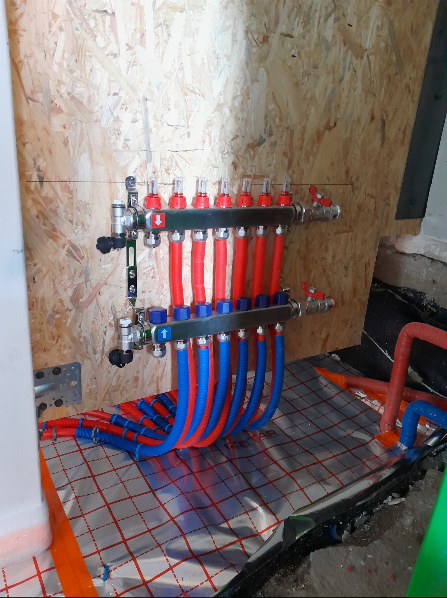 Rozdzielacz ogrzewania podłogowego z czerwonymi i niebieskimi rurami na folii izolacyjnej, zamontowany na ścianie z płyty OSB. Widok na instalację w trakcie montażu.