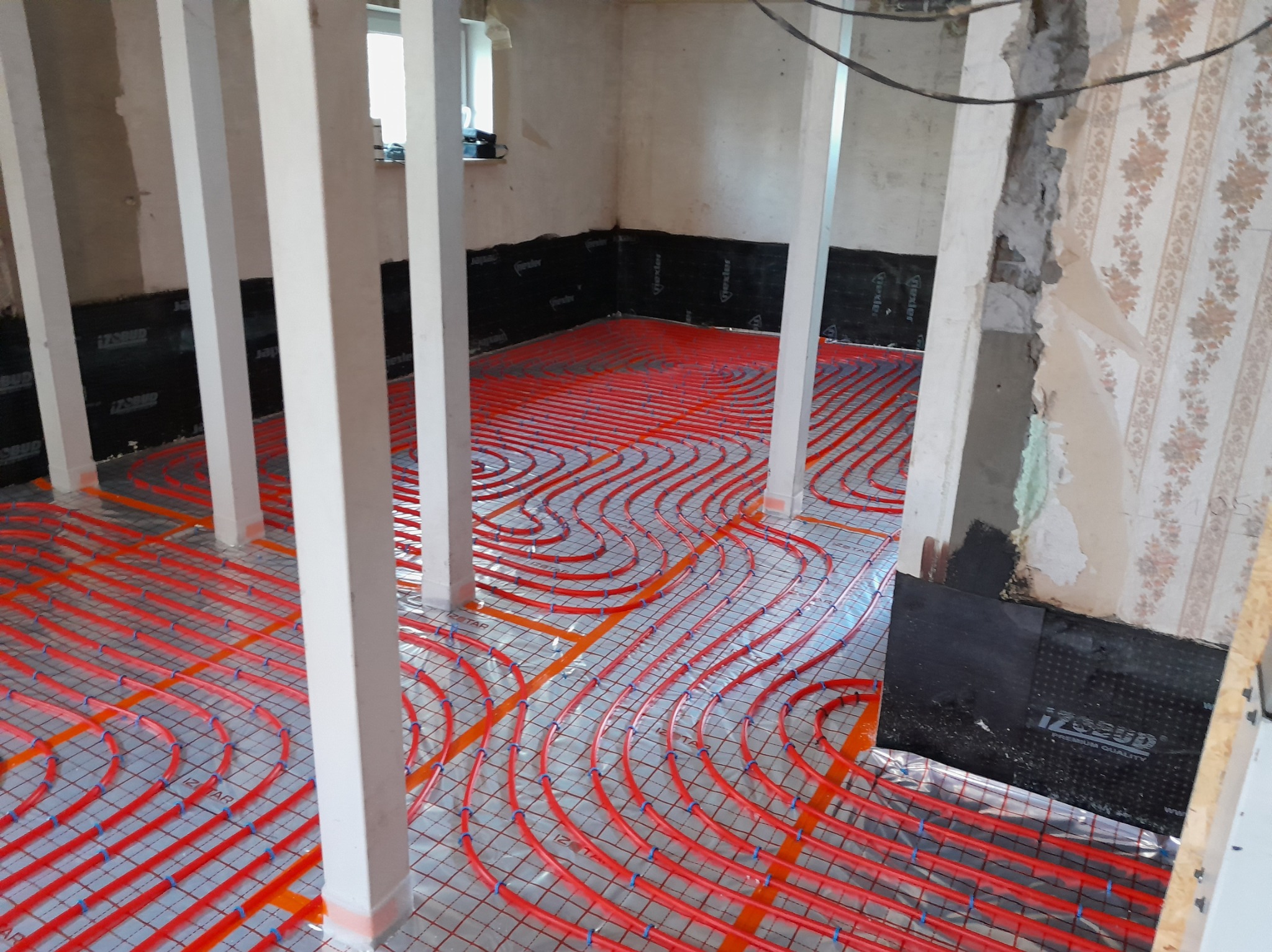 Czerwone rury ogrzewania podłogowego ułożone na folii, widoczne białe słupy konstrukcyjne i fragment ściany w trakcie remontu.