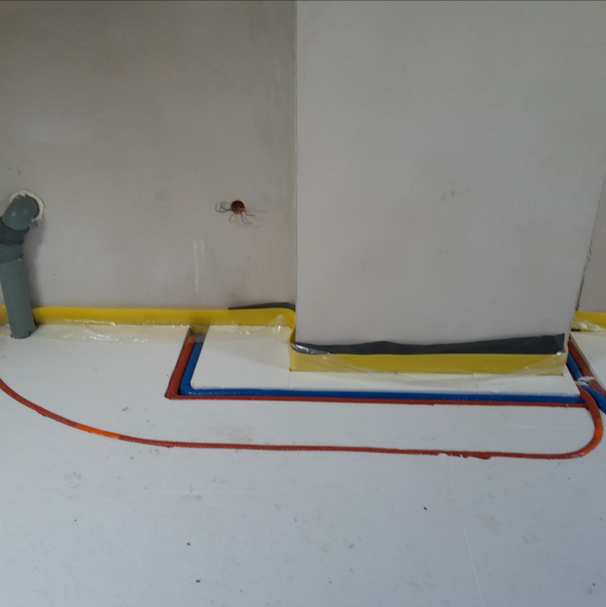 Montaż ogrzewania podłogowego: czerwone i niebieskie rurki na białym styropianie, żółta taśma brzegowa przy ścianie. Widoczny fragment instalacji wodnej.