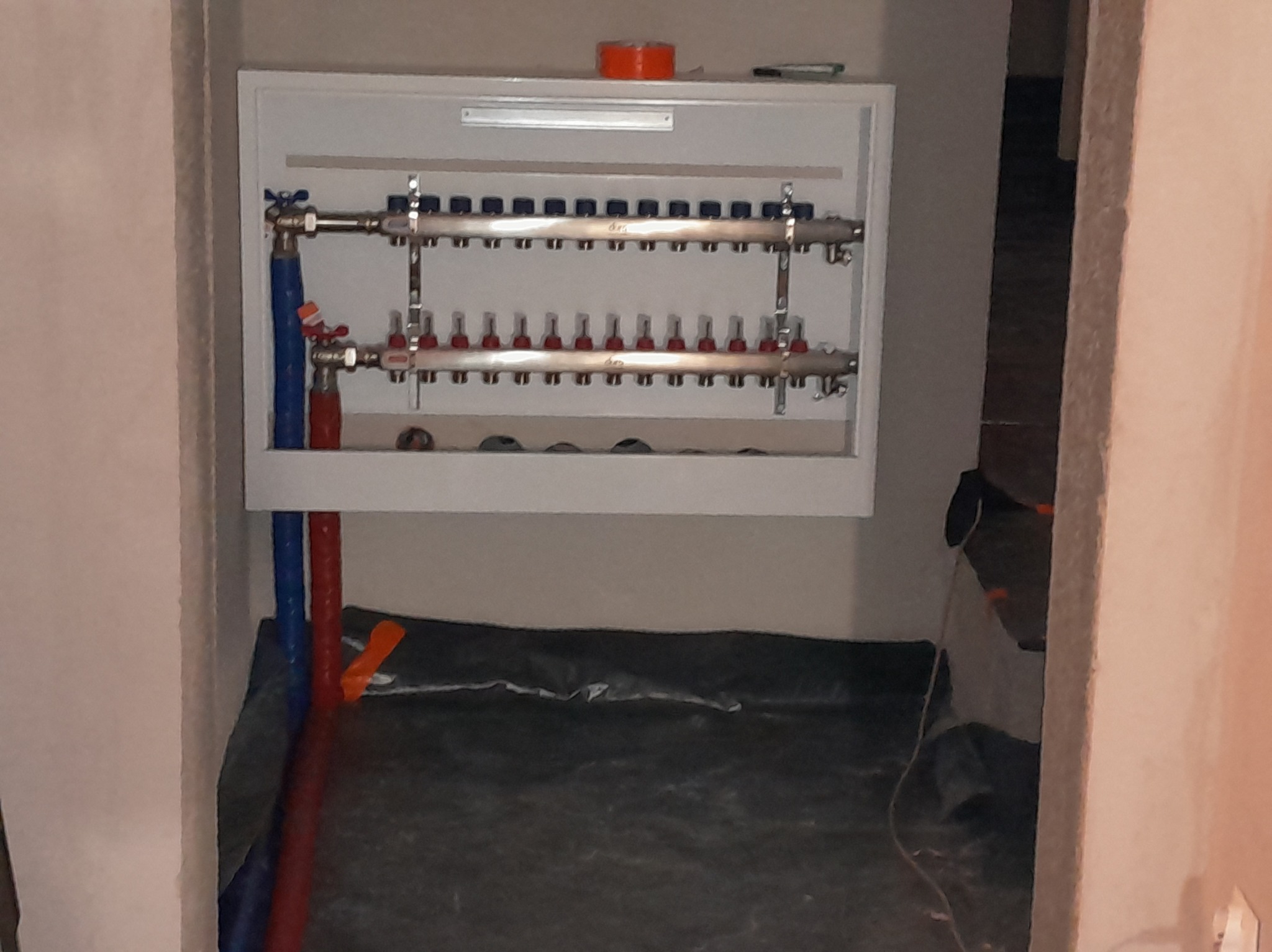 Rozdzielacz ogrzewania podłogowego w białej szafce, z niebieskimi i czerwonymi rurami, w surowym wnętrzu. Na szafce pomarańczowa taśma.