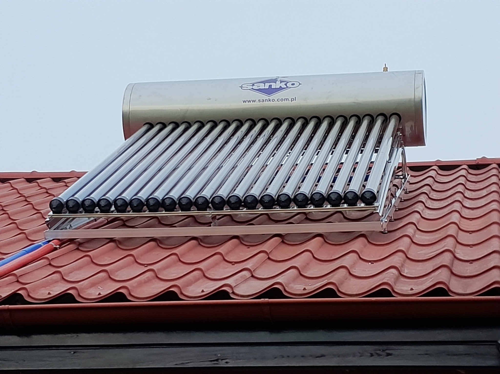 Kolektor słoneczny Sanko na czerwonym dachu, widok z dołu. Srebrny zbiornik z niebieskim logo i rzędem szklanych rur. Instalacja solarna na tle jasnego nieba.