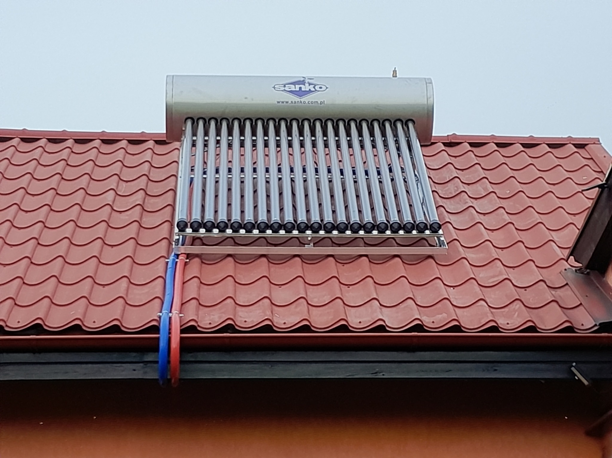 Kolektor słoneczny Sanko z rurami na czerwonym dachu. Widoczne mocowania i instalacja hydrauliczna. Szare niebo w tle.