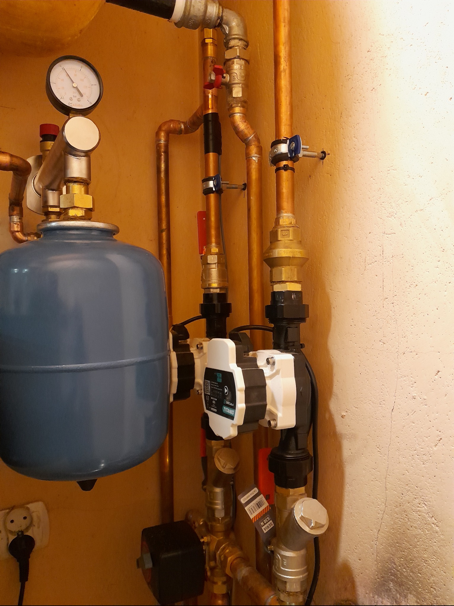 Złożona instalacja hydrauliczna z miedzianymi rurami, manometrem i pompą obiegową Titanio, zamontowana na ścianie o kremowej fakturze. Widoczne złącza i zawory.