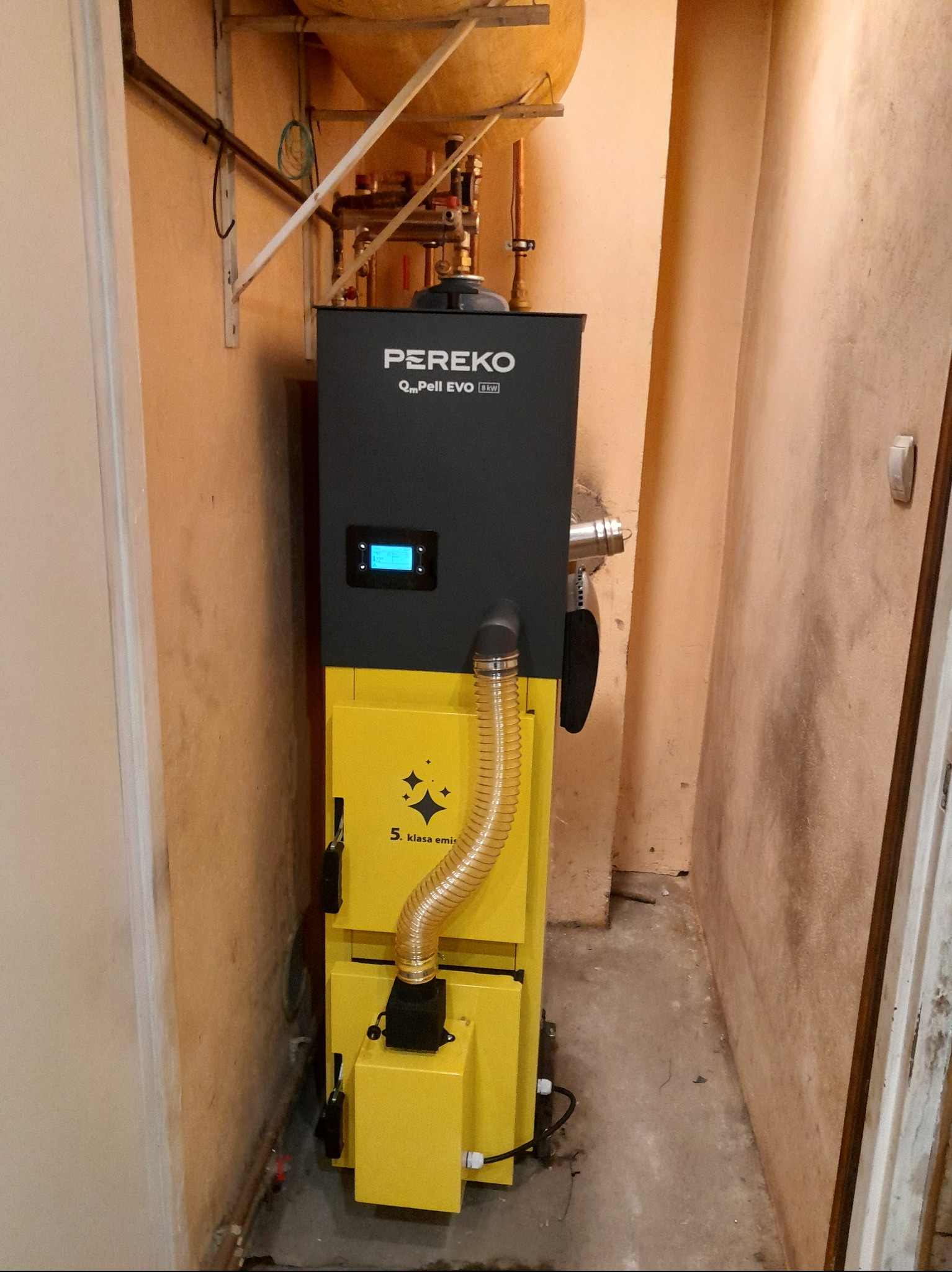 Żółto-czarny piec Pereko Q-Pell EVO 8kW w kotłowni, widoczny wyświetlacz i rura spalinowa. Piec ustawiony przy ścianie, widoczne rury instalacji grzewczej.