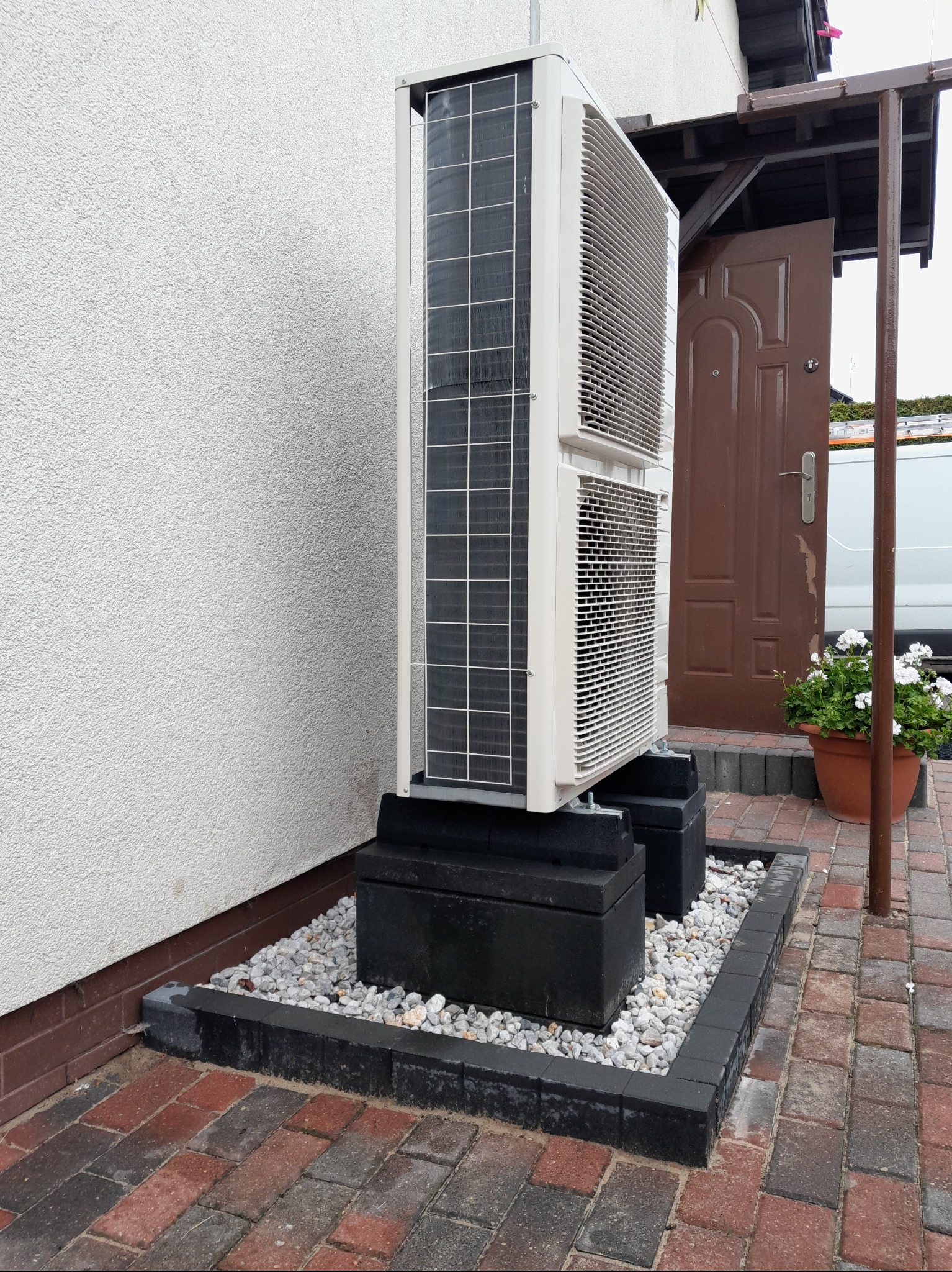 Biała pompa ciepła typu split, ustawiona na czarnych betonowych blokach, otoczona białym żwirem i kostką brukową, na tle białej ściany budynku i brązowych drzwi.