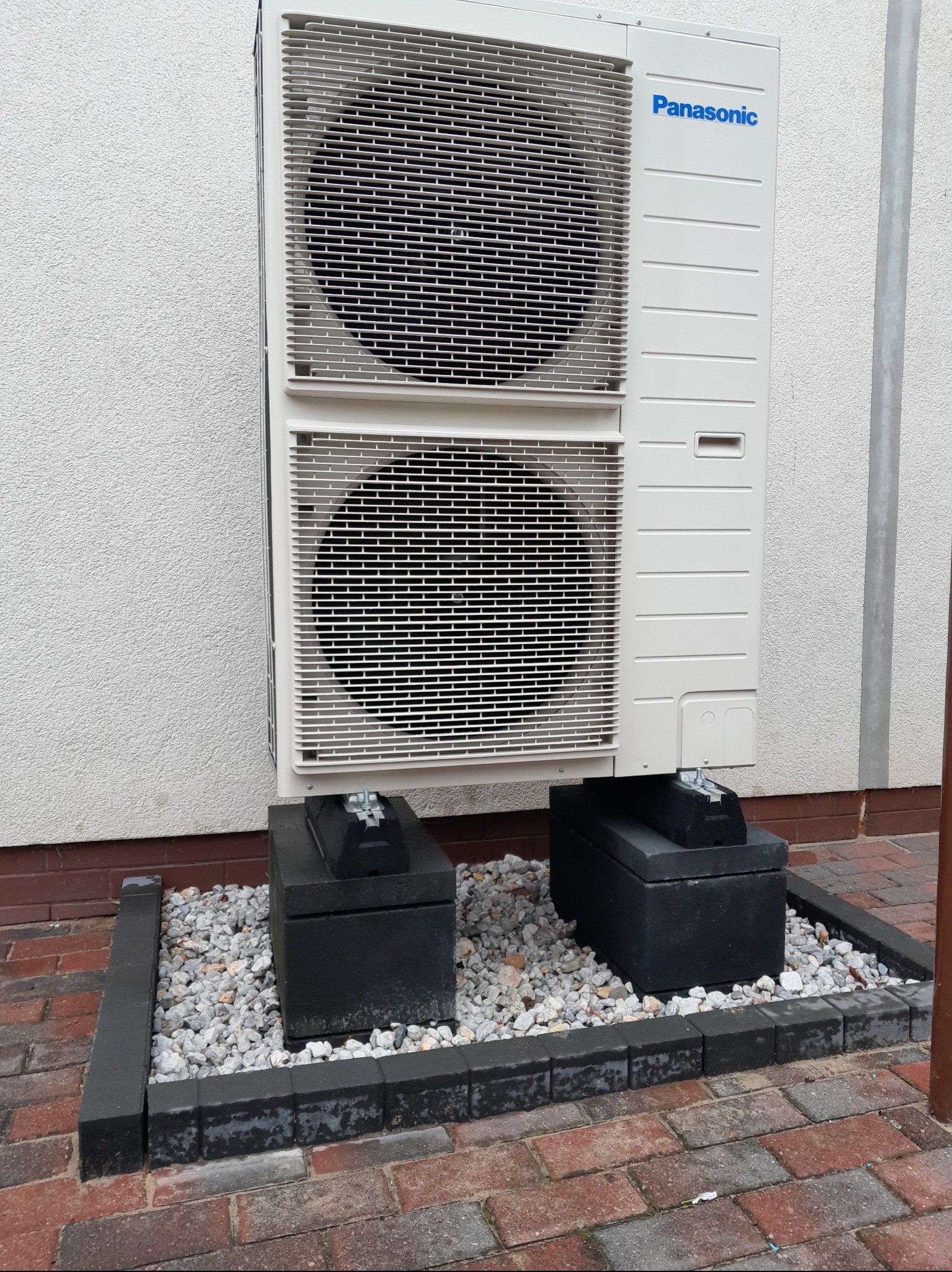 Biała, dwuczęściowa pompa ciepła Panasonic zamontowana na betonowych blokach, otoczona kamieniami i obrzeżem z kostki, przy ścianie budynku.