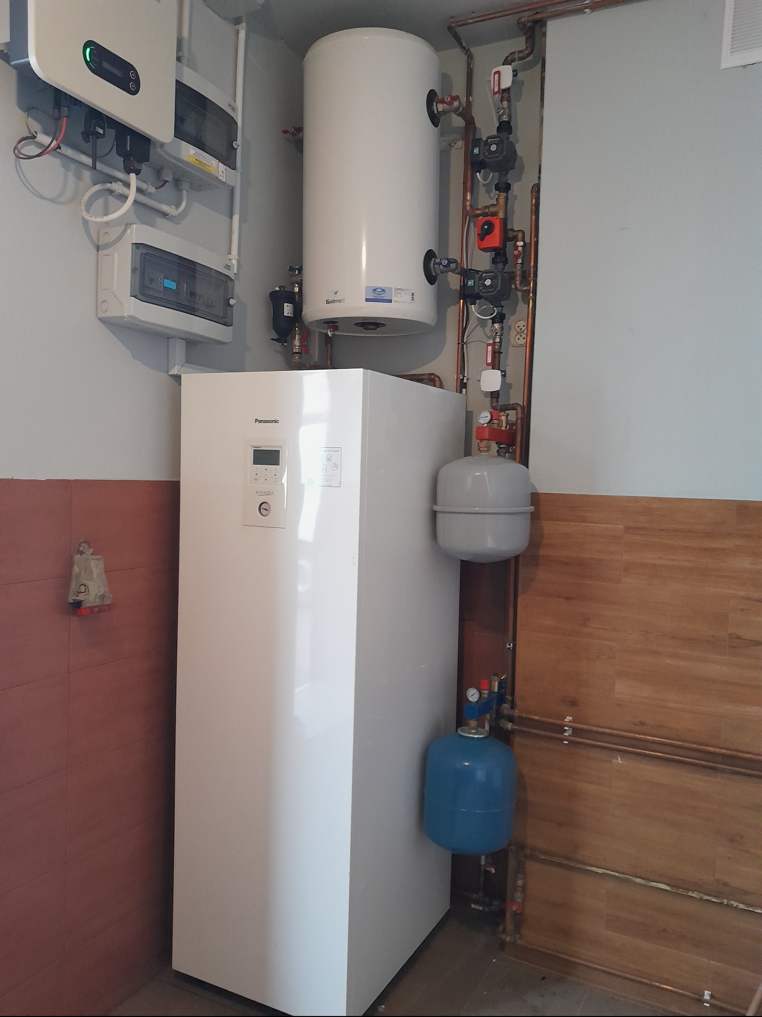 Biała pompa ciepła Panasonic Aquarea w kotłowni, z widocznymi elementami instalacji, zbiornikiem i orurowaniem. Na ścianie panele sterowania i zabezpieczeń.