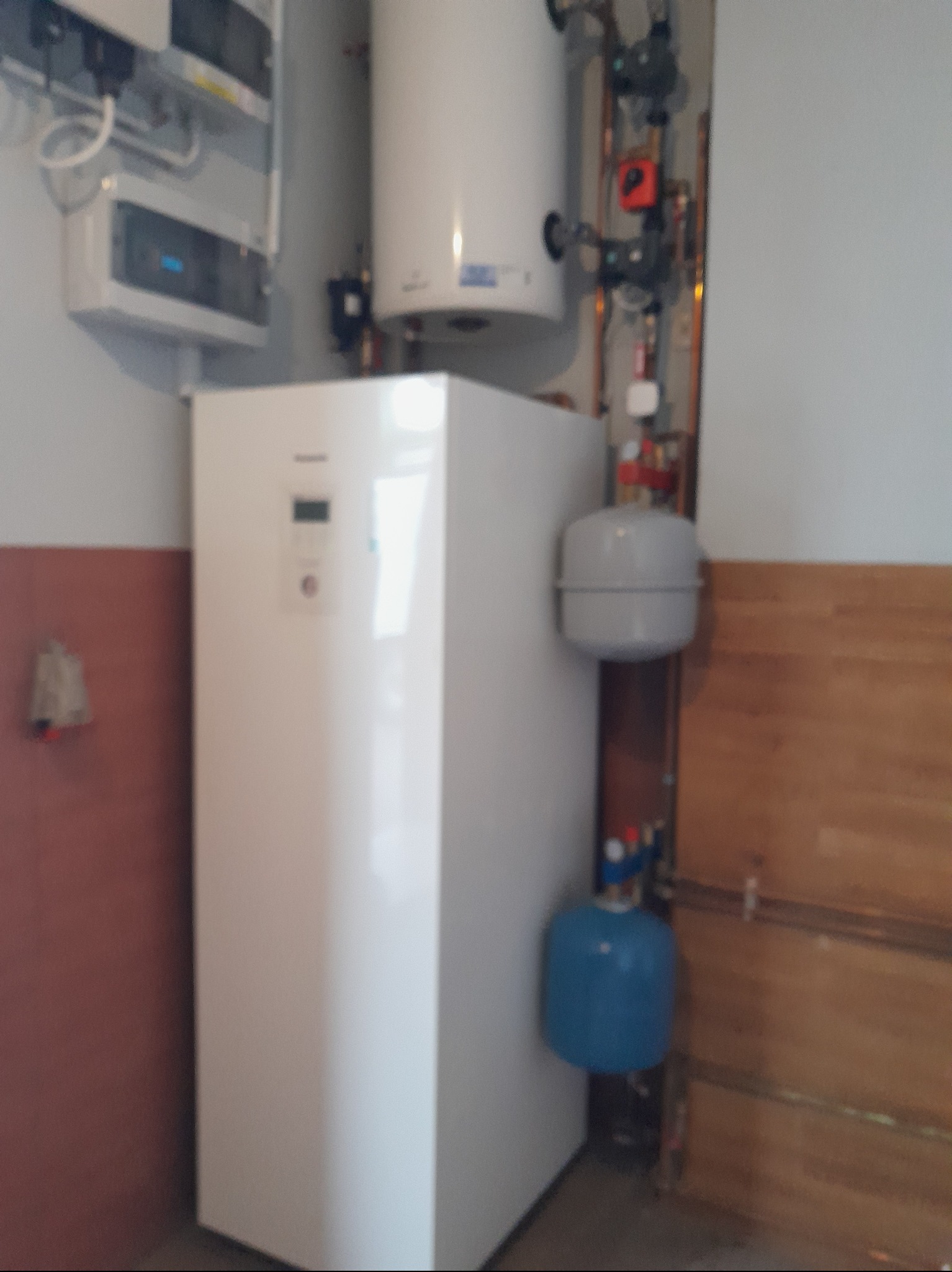 Biała pompa ciepła Panasonic z zbiornikiem, widoczne rury i zawory. Elementy instalacji grzewczej na tle ściany w dwóch kolorach. Ujęcie z bliska, lekko zamazane.