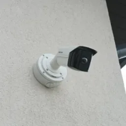 Biała kamera monitoringu wizyjnego marki Hikvision, zamontowana na jasnej, chropowatej ścianie budynku, z widocznym fragmentem rynny.