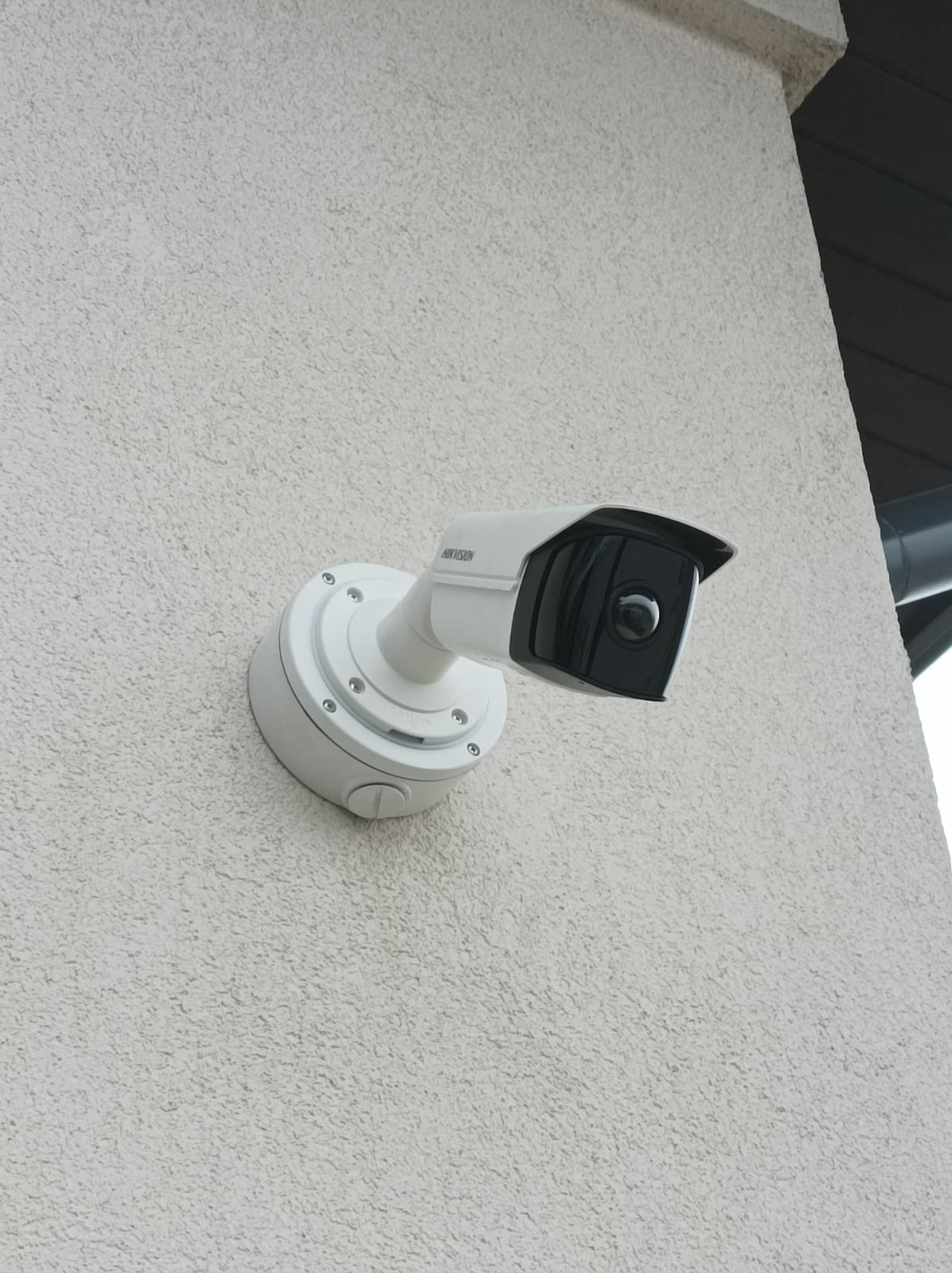 Biała kamera monitoringu wizyjnego marki Hikvision, zamontowana na jasnej, chropowatej ścianie budynku, z widocznym fragmentem rynny.