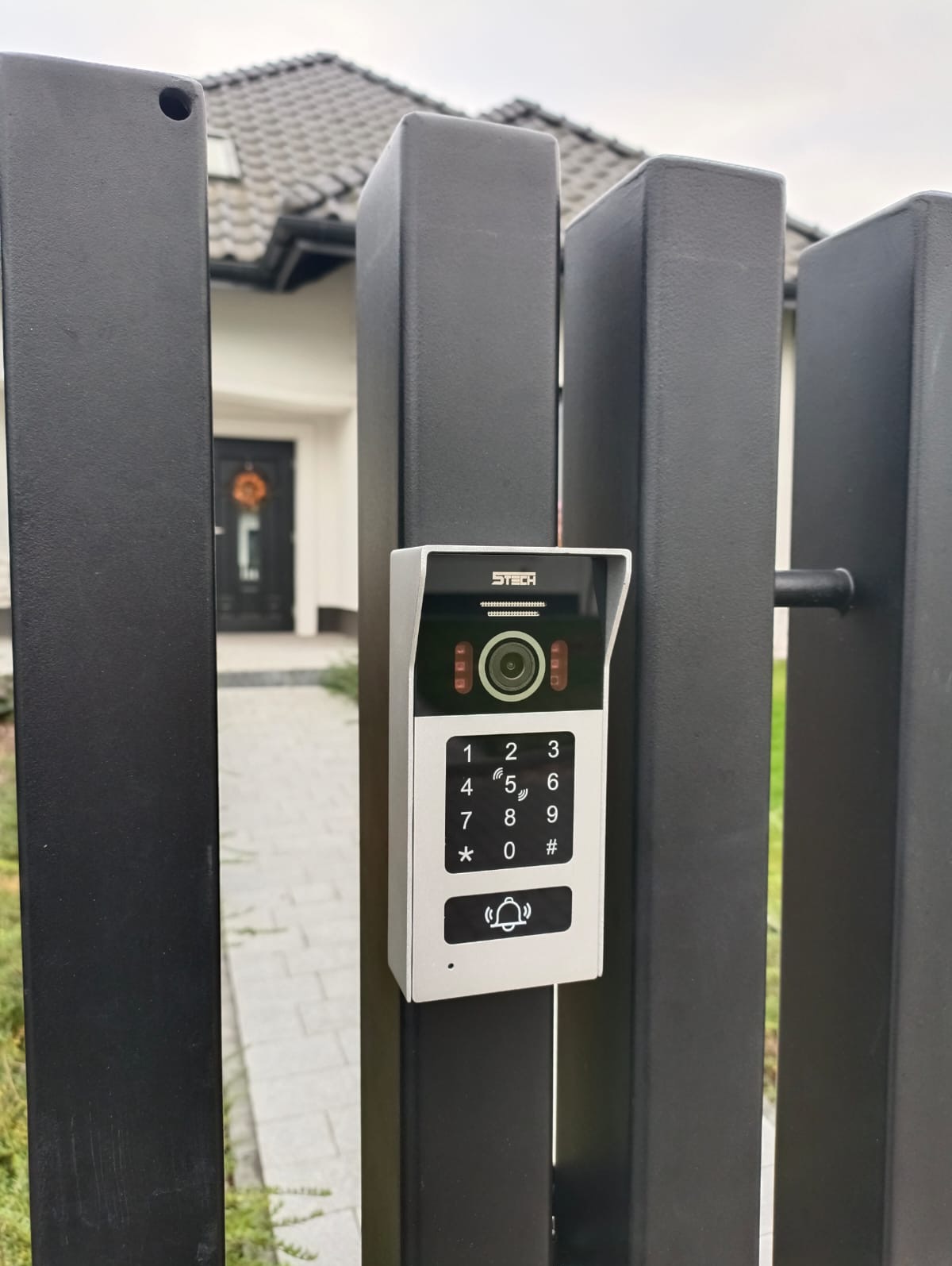 Szary wideodomofon 5Tech z klawiaturą numeryczną i przyciskiem dzwonka, zamontowany na czarnym, metalowym słupku ogrodzenia posesji, z widocznym fragmentem domu w tle.