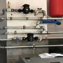 Rozbudowana instalacja grzewcza z pompami obiegowymi, zaworami i rurami na ścianie, widoczny zbiornik wyrównawczy i otwarty podręcznik techniczny na blacie.