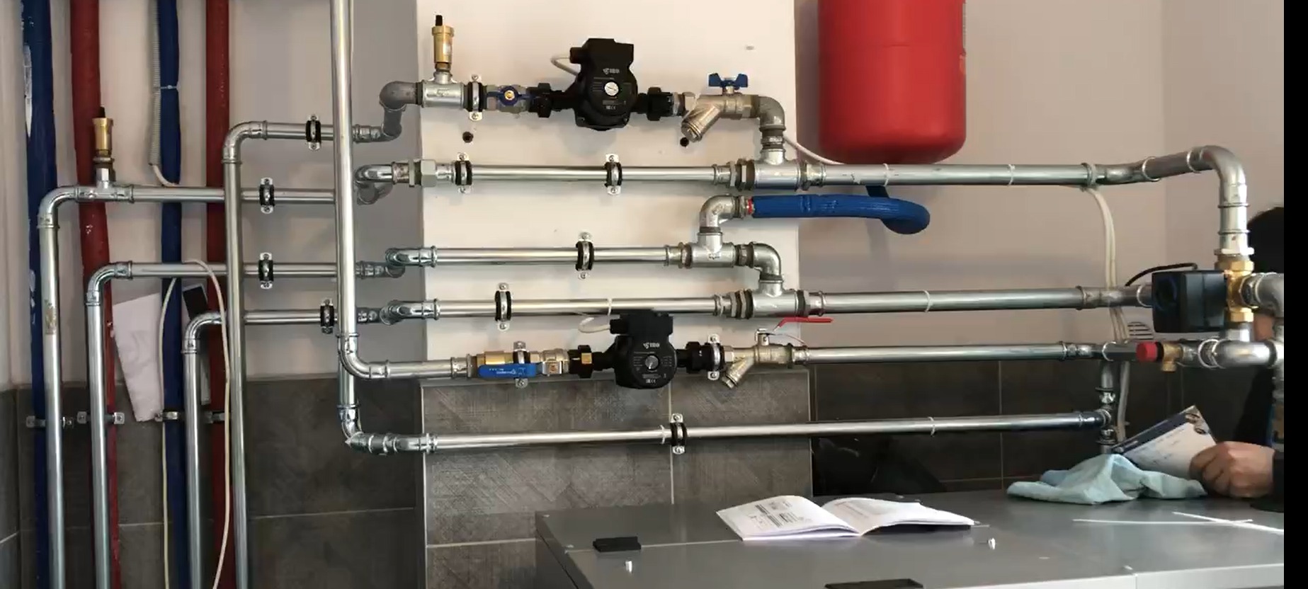 Rozbudowana instalacja grzewcza z pompami obiegowymi, zaworami i rurami na ścianie, widoczny zbiornik wyrównawczy i otwarty podręcznik techniczny na blacie.