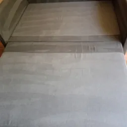 Sofa w rozłożonej formie z widocznymi śladami czyszczenia na tapicerce, ukazującymi różnicę w odcieniach szarości.