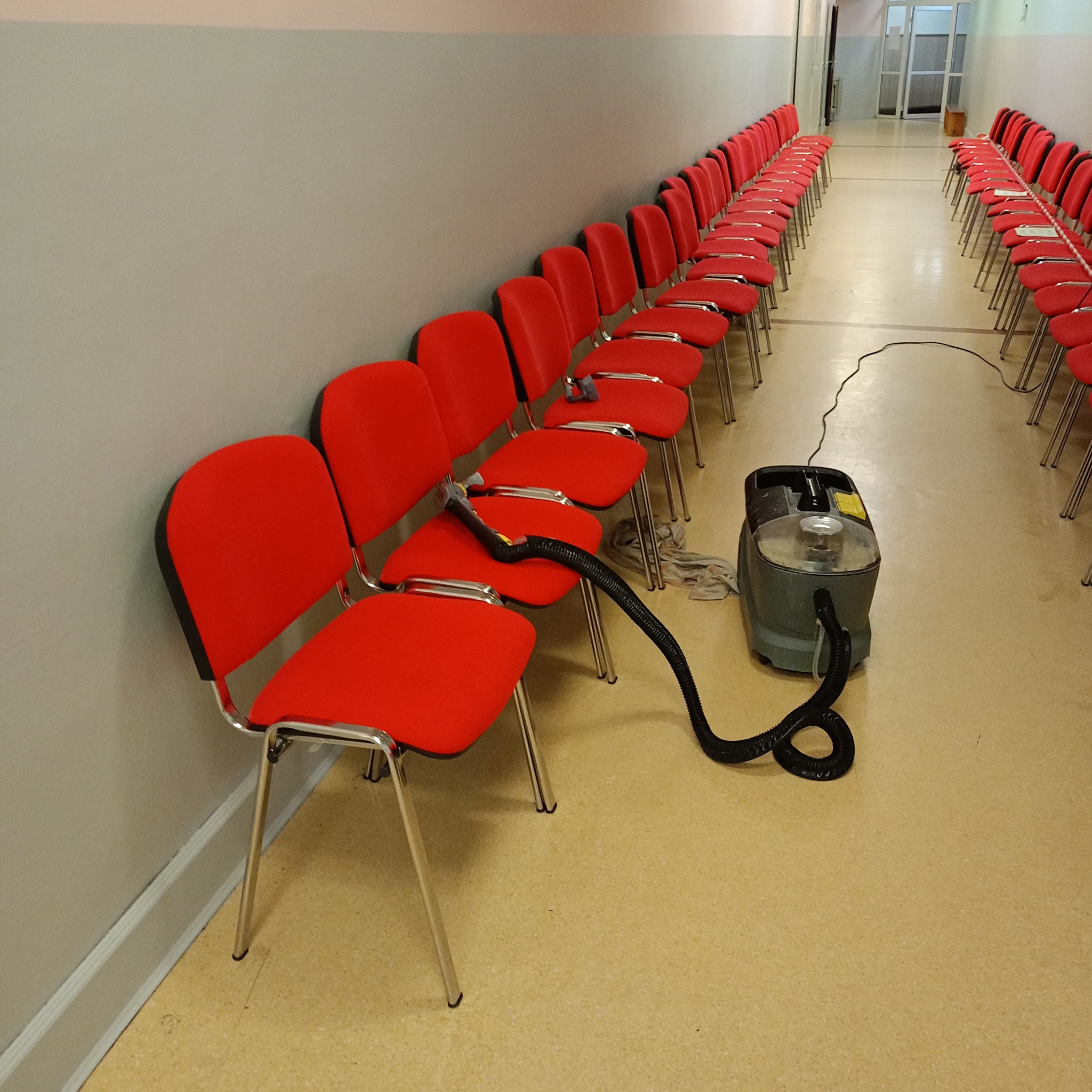Rząd czerwonych krzeseł z tapicerką, czyszczonych za pomocą odkurzacza piorącego z czarnym wężem, stojącego na beżowej podłodze wzdłuż szarej ściany.
