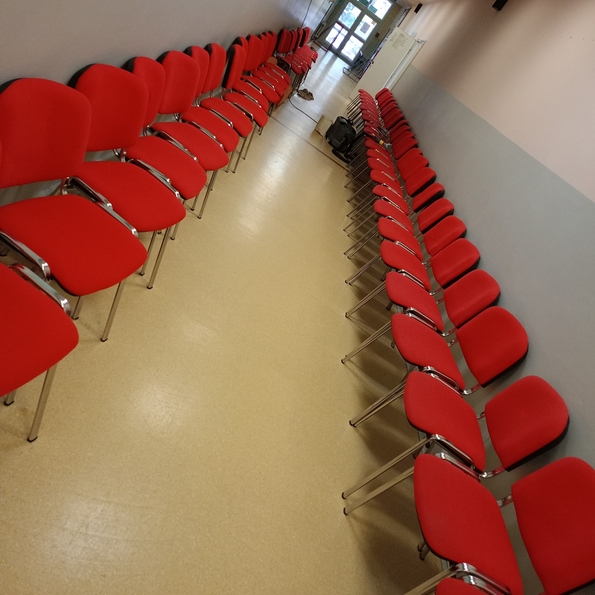 Rząd czerwonych krzeseł z chromowanymi nogami ustawionych wzdłuż jasnej ściany w pomieszczeniu z beżową podłogą, perspektywa zbieżna w kierunku drzwi na końcu korytarza.