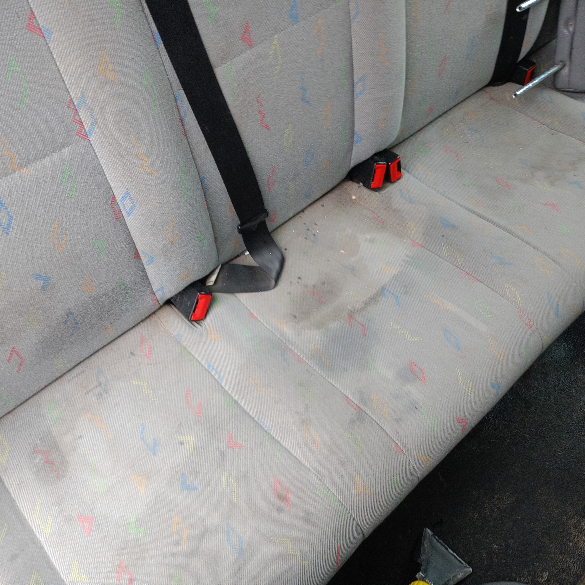 Brudna, szara tapicerka samochodowa z kolorowymi wzorami i czarnym pasem bezpieczeństwa, widoczne plamy przed czyszczeniem.