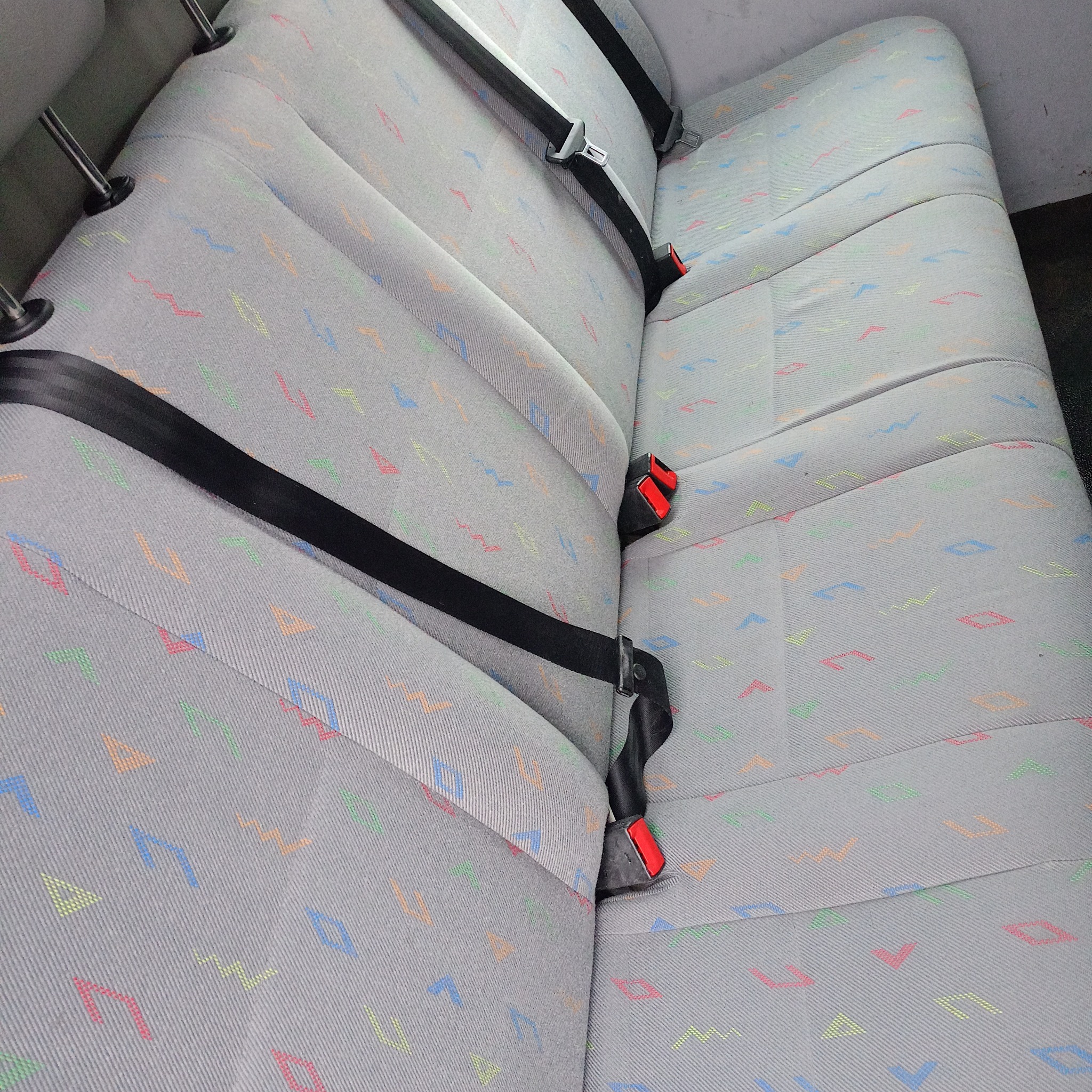 Szare, tekstylne siedzenia samochodowe z kolorowym wzorem geometrycznym i czarnymi pasami bezpieczeństwa.