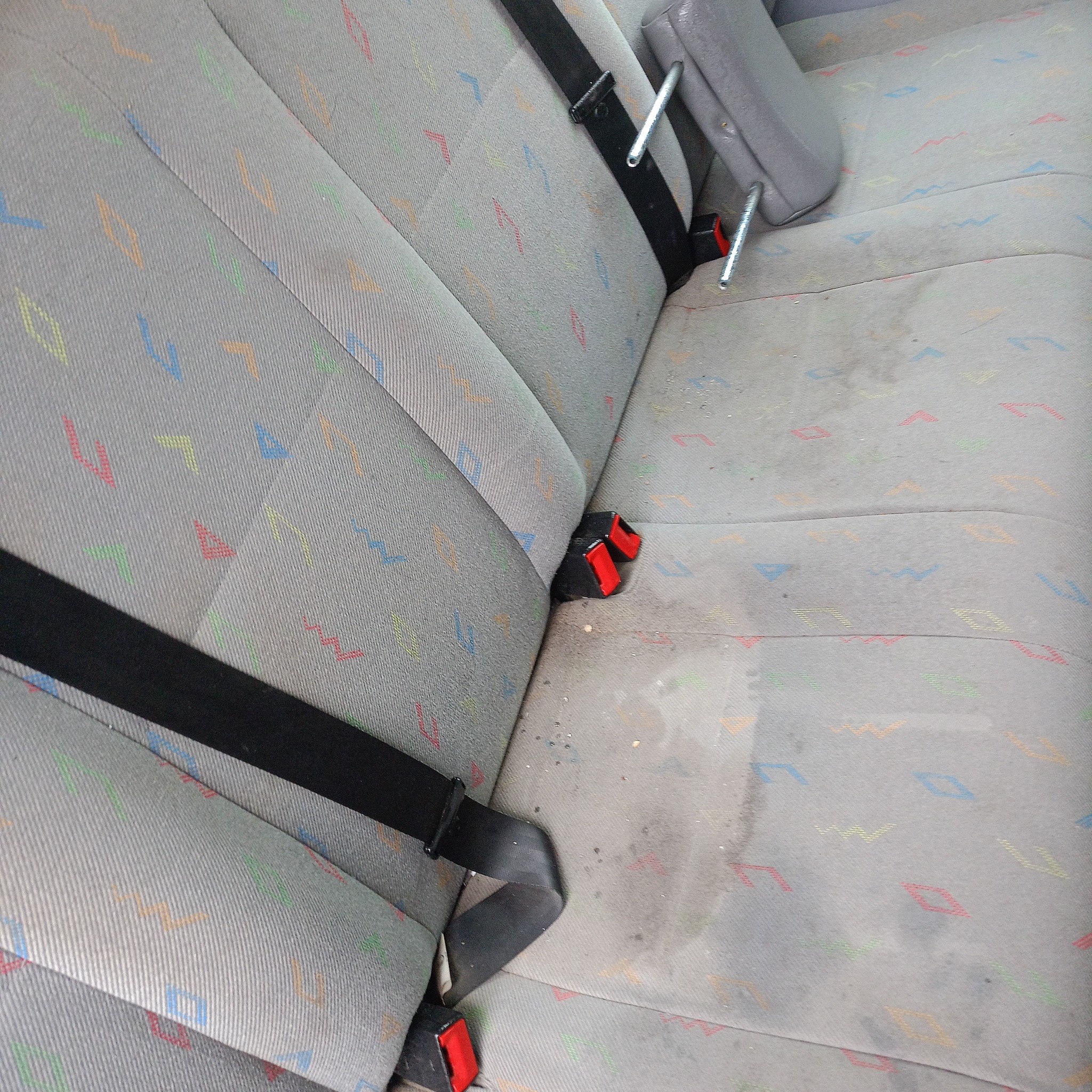 Szara, zabrudzona tylna kanapa samochodowa z kolorowym wzorem geometrycznym i czarnymi pasami bezpieczeństwa.