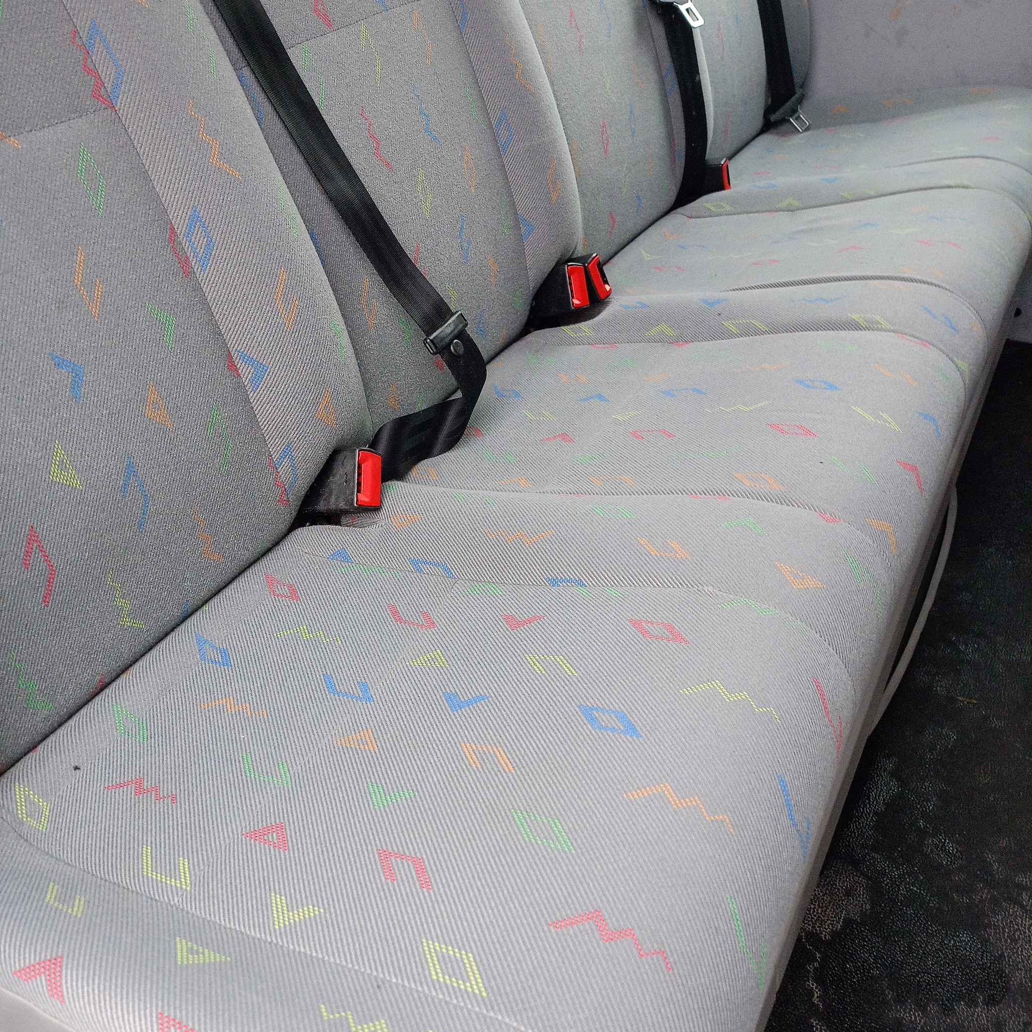 Szara tapicerka samochodowa z kolorowym wzorem geometrycznym, widoczne pasy bezpieczeństwa z czerwonymi elementami.