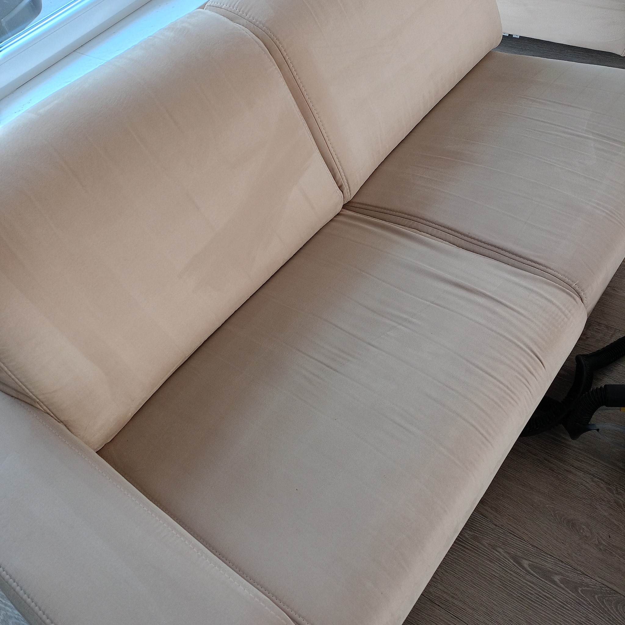 Beżowa kanapa z widoczną różnicą w odcieniu po czyszczeniu, fragment węża odkurzacza w prawym dolnym rogu.