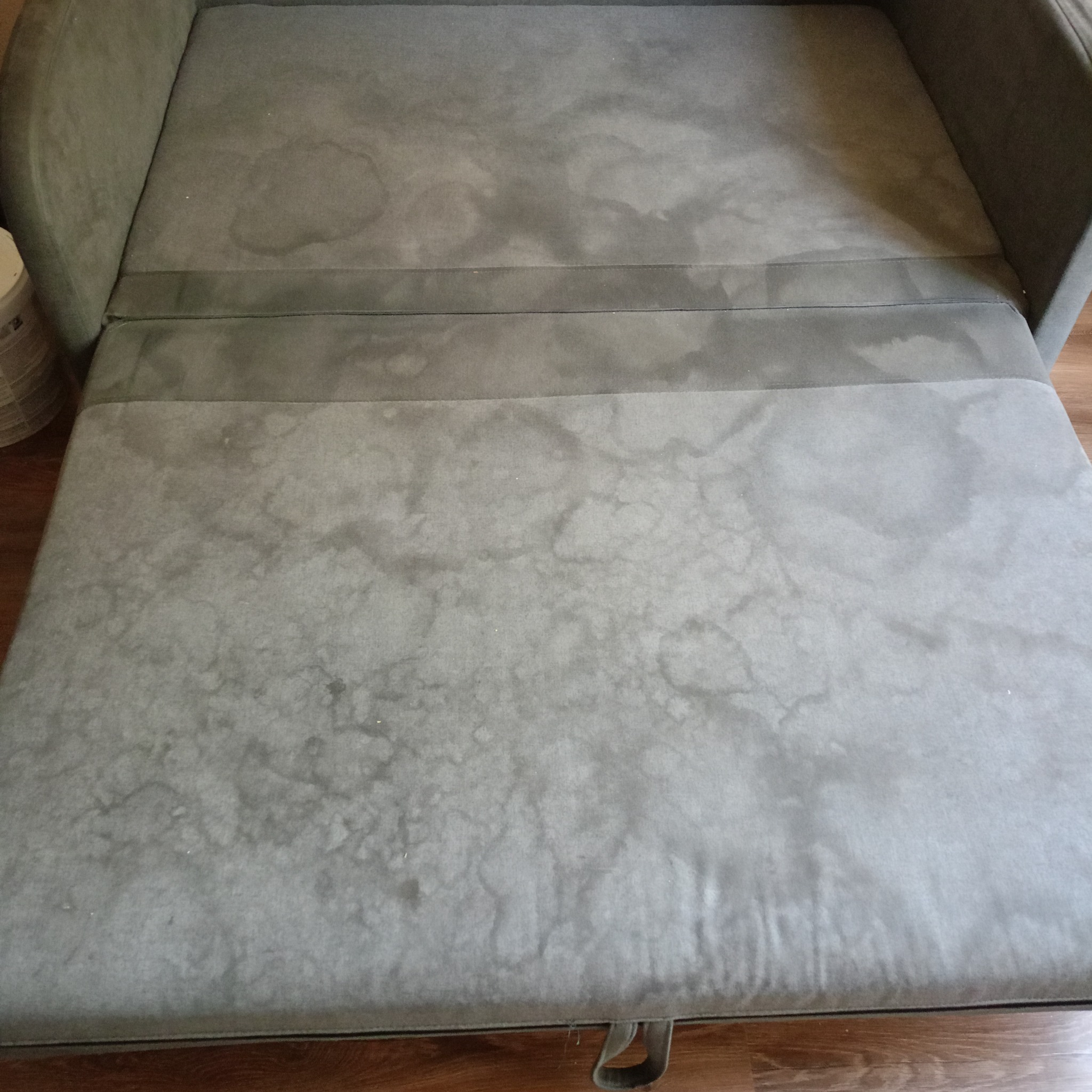 Rozłożona szara sofa z widocznymi plamami na tapicerce, leżąca na drewnianej podłodze.