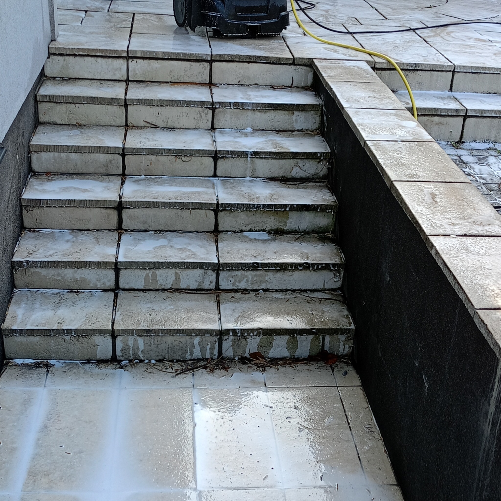 Kamienne schody i taras w trakcie mycia pod ciśnieniem. Widoczna woda i resztki brudu. Czarny sprzęt do mycia ciśnieniowego na górze schodów.