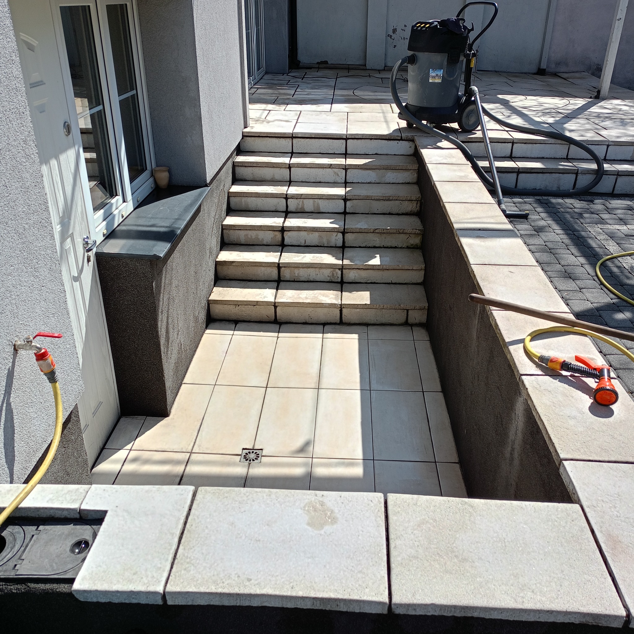 Czyszczenie kostki brukowej na schodach i tarasie przy użyciu odkurzacza przemysłowego. Widoczny kran z wężem i dyszą. Jasne płytki tarasowe.