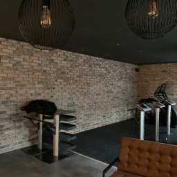 Solid-Art - Surowe ceglane ściany z dekoracyjnymi lampami wiszącymi, brązowa sofa i sprzęt do ćwiczeń w tle, tworzące nowoczesną przestrzeń.