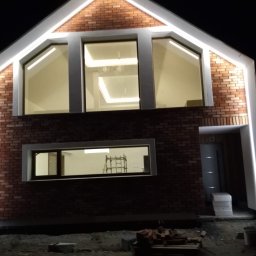 Solid-Art - Dwukondygnacyjny budynek z cegły z oknami oświetlonymi od wewnątrz i na zewnątrz białym światłem LED, widoczne rusztowanie wewnątrz budynku przez okno na parterze.