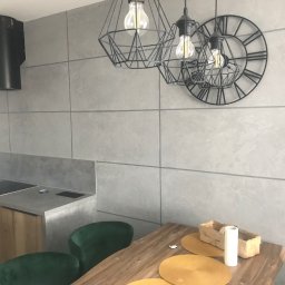 Solid-Art - Szara kuchnia z wyspą, zielonymi krzesłami, geometrycznymi lampami i zegarem na ścianie z szarych płyt.