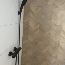 Solid-Art - Czarny zestaw prysznicowy zamontowany na białej, kafelkowanej ścianie, z podłogą wyłożoną parkietem w jodełkę w tle.