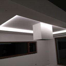 Solid-Art - Nowoczesny sufit napinany z oświetleniem LED w kształcie prostokąta, z centralnym elementem dekoracyjnym w formie białego prostopadłościanu, widoczne okno z ciemną szybą.