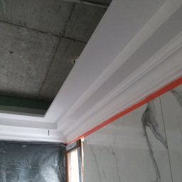 Solid-Art - Montaż ozdobnych listew przysufitowych w pomieszczeniu z surowym betonowym stropem i marmurowymi płytkami ściennymi, zabezpieczonym folią oknem.