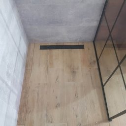 Solid-Art - Ułożone płytki podłogowe imitujące drewno w kabinie prysznicowej z czarnym odpływem liniowym i szklaną ścianką. Widoczne również szare płytki ścienne.