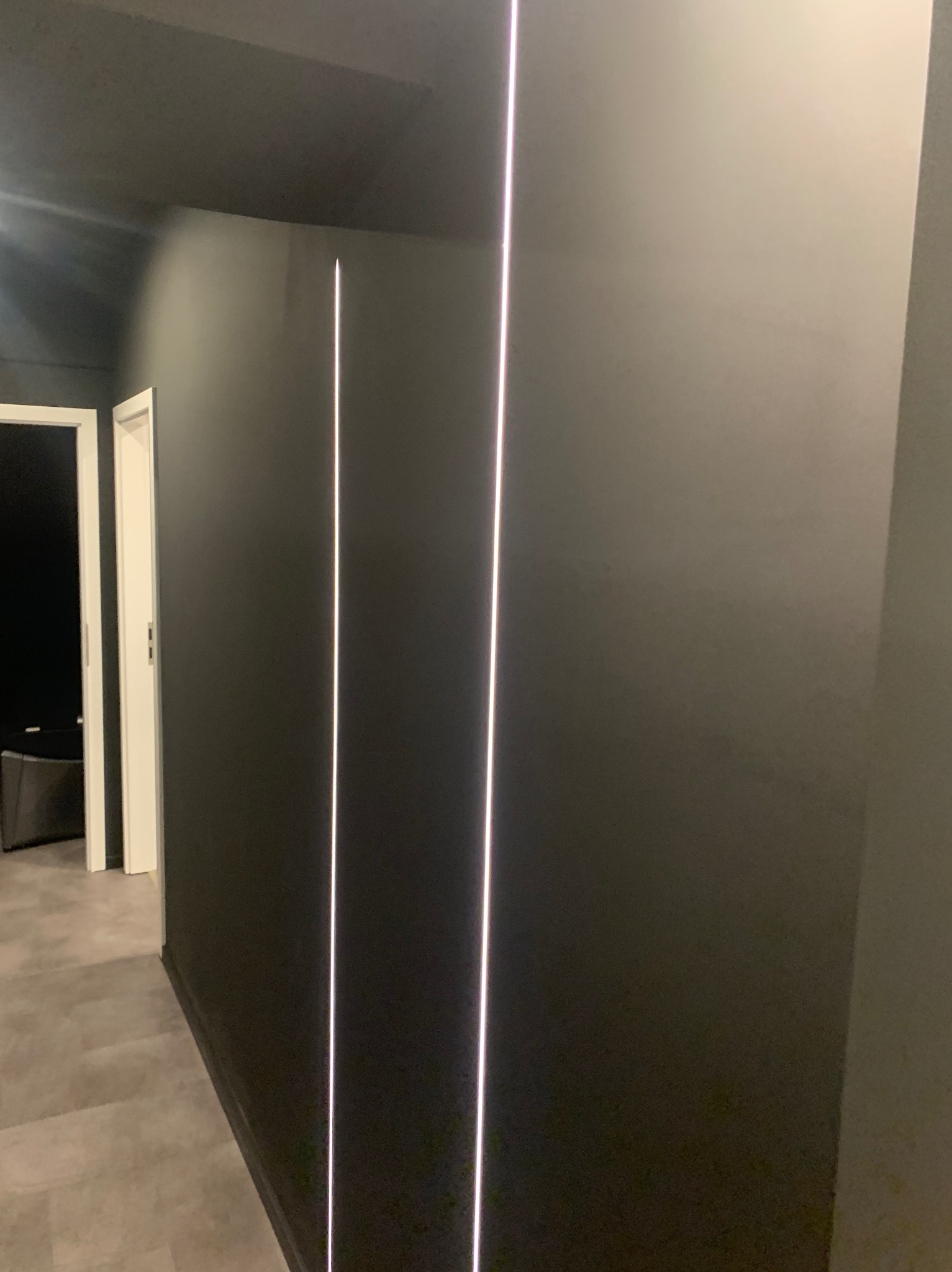 Szara ściana z pionowymi wbudowanymi paskami LED, widok na korytarz z szarą podłogą i białymi drzwiami.